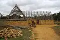 Photographie montrant un bâtiment en pierres recouvert d'une charpente formée de troncs, avec un artisan et deux personnes l'entourant.