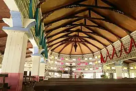 Photo en couleur montrant les piliers et le toit en voûte de la basilique. De nombreuses ouvertures laissent passer la lumière. Une croix est visible en fond.