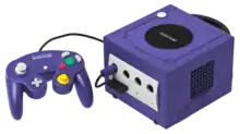 Console de jeux vidéo cubique de couleur violette, avec une manette filaire de même couleur équipée de boutons multicolores.