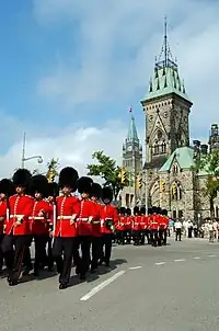 Photo en couleurs de militaires portant une tunique rouge et une bonnet à poil noir défilant sur une rue devant un bâtiment comprenant une tour