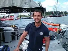 Le navigateur en buste, polo bleu marine, à l'arrière de son bateau au ponton.
