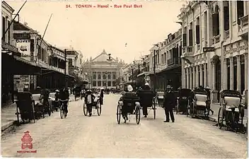 Photo ancienne montrant une rue dans laquelle circulent des pousse-pousses, avec en arrière-plan un grand bâtiment.
