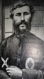 Photo d'un prêtre, barbu, un Sacré-Cœur sur la poitrine et un crucifix à la main, devant une cloison