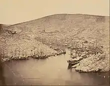 Photographie d'une baie étroite bordée de collines rocailleuses descendant à pic. La baie est presque entièrement remplie de navires à voiles.