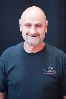 Photographie couleur d’un homme, de tronc, chauve, souriant, avec une barbe de 3&nbsp;jours blanche et un t-shirt bleu marine