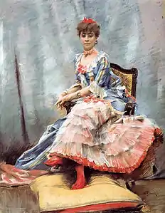 Portrait de Laure Hayman, assise de trois quarts sur un fauteuil, les deux mains reposant sur accoudoir de droite et les pieds gainés de bas rouges et chaussés de même couleur reposent sur un gros coussin bicolore, jaune paille au-dessus, rouge en-dessous. Le bleu, le rose et le rouge dominent dans cette œuvre, donnant une impression de légèreté, de jeunesse, de luminosité. Le rouge se retrouve dans le nœud qui surmonte des cheveux bouclés serrés en chignon, dans celui qui enserre le cou, dans celui qui orne la pointe du décolleté bordé d’un volant de la robe bleue et jaune dont les manches arrêtées à hauteur des coudes se terminent elles aussi par un volant, et dont la jupe, ouverte sur le devant, s’achève en traîne. Du rouge encore pour la jupe de fond recouverte de cinq volants de tulle ou de mousseline qui donnent par transparence un effet rose incarnat rappelant celui des lèvres et des joues fardées. Un bleu et un rose plus légers forment aussi la trame de la draperie en arrière fond.