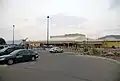 A local parking lot with a number of automobiles using the service; there is also a building of an airport, which contains its terminal.