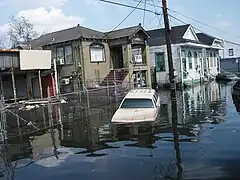 Photographie d'une rue inondée avec une voiture dans l'eau et plusieurs maisons aux fondations recouvertes.