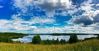 Un lac bleu cristallin par ciel bleu avec des nuages, dans un paysage entouré de forêts de taïga.