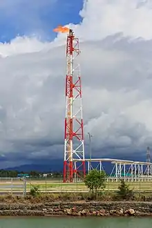 Des installations métalliques et des conduites blanches sont construites sur un terrain couvert d'herbe. En leur centre se trouve une torchère enflammée, dont les couleurs rouges et blanches contrastent avec le ciel aux nuages sombres. Au premier plan, des grillages barrent l'accès au site, et une montagne ainsi que des arbres sont visibles à l'arrière-plan.