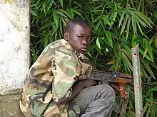 Photographie couleur d'un enfant accroupi tenant un fusil automatique.