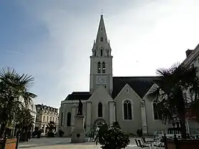 Photographie d'une place ornée de palmiers en bacs. Au centre, statue de bronze sur un haut piédestal. Au second plan une église : le chœur coté gauche, le clocher au centre (à la croisée des transepts).
