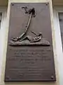 Photographie depuis le bas d'une plaque en métal sombre ; la partie supérieure étant une sculpture en relief, la partie inférieur un texte en hongrois sur plusieurs lignes