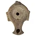 Cliché couleur. Objet en terre cuite vu de dessus avec un réservoir, deux ailerons et deux orifices laissant passer la mèche et son réglage.