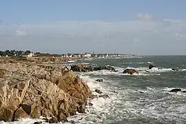 Côte rocheuse peu élevée contre laquelle des vagues viennent se briser, sur fond de ciel bleu.