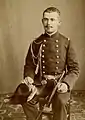Photographie en noir et blanc d'un homme assis, portant une tenue de bordache, le poignard à la ceinture et le bicorne à la main.