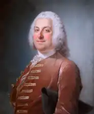 Portrait en buste d’un homme portant perruque, avec veste de velours rouge et tricorne sous le bras gauche.