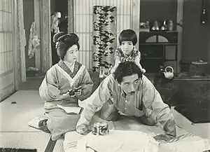 Une femme à genou (Kinuyo Tanaka) tient dans ses mains un porte-monnaie ouvert et regarde sa fille (Mitsuko Ichimura) assise à califourchon sur le dos de son père (Atsushi Watanabe) et qui s’agrippe à ses cheveux (image en noir et blanc).