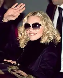 Une femme blonde habillée d'une veste mauve, portant des lunettes de soleil et saluant la foule