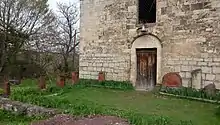 Photographie couleur de la façade d'un bâtiment en pierre de taille, dont l'entrée est fermée par une porte en bois. Au premier plan, des khatchkars sont disposés sur la pelouse.