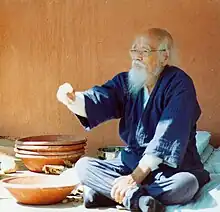 C'est une photographie en couleur de Fukuoka assis devant des récipients en céramique, il a un âge avancé.