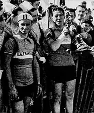 Photo en noir et blanc montrant deux hommes en tenue de cycliste se tenant devant des spectatrices portant des coiffes