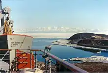 Proue d'un bateau vue depuis une plate-forme, avec une péninsule occupée par une cabane et, au-delà d'une mer couverte de glace, une chaîne montagneuse.