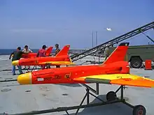 Trois drones rouges fuselés avec rampe sur pont de porte-avion. Les ailes delta et les nez sont jaunes (3 × 2,5 m env.).