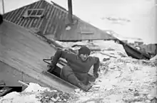 Photographie d'un homme sortant par une ouverture dans le toit d'un petit bâtiment en bois.