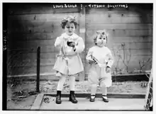 photo en noir et blanc de deux enfants debout habillés en blanc, l'un brun, l'autre, plus petit, blond
