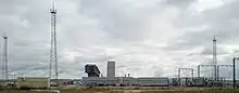 Des installations industrielles comportant plusieurs tours et structures métalliques, à droite et à gauche de l'image et des bâtiments, devant un ciel nuageux.