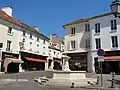 Fontaine en pierre blanche, à quatre jets, devant des immeubles anciens à façade blanche