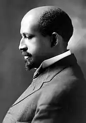 Photo ancienne noir et blanc de profil d'un homme noir en costume avec une chemise blanche penchant légèrement la tête en avant, le front dégarni, avec un bouc et une moustache courte.
