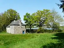 Bâtiment et pierre grise et toit d'ardoise noire, sur une zone herbeuse au milieu d'arbres. À l'arrière on aperçoit un plan d'eau.
