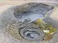 Une mare de boue à Krafla Islande. Notez qu'il s'agit probablement de la même que celle montrée plus en haut, mais prise plus tard, la cavité du bas est d'ailleurs presque tarie et des dépôts de soufre sont clairement visibles autour de la source de jaillissement.