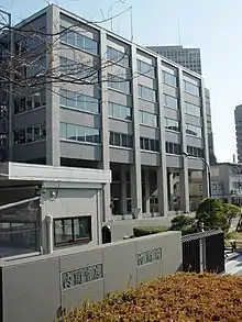 Immeuble moderne en pierre grise de cinq étages, les quatre étages supérieurs s'avançant plus avant, vu de 3/4 gauche. Au premier plan, un mur bas construit avec les mêmes pierres sur lequel est inscrit bureau du cabinet en japonais et un portail noir. À gauche, derrière le mur, se trouve également une cabane de gardien.