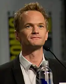 L'acteur Neil Patrick Harris, en costume cravate et assis derrière un micro dans le cadre d'une interview, est blond aux yeux bleus, et a également des rides visibles sur le front et un début de calvitie près de ses tempes.