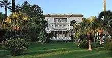 Vue d'un jardin avec palmiers au fond duquel une bâtisse blanche imposante à toit plat et entrée à colonnes se détache.