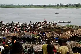 Photographie couleur montrant une foule et des pirogues au bord d'un large fleuve.
