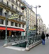 Photographie en couleurs d'une bouche de métro, avec ferrures vert de gris et perspective diagonale d'une rue se prolongeant vers le côté droit de l'image