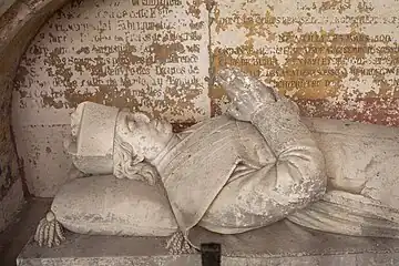 Gisant de pierre représentant un homme, en costume ecclésiastique, joignant les mains.