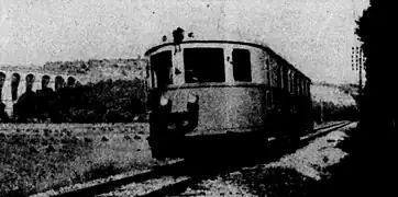 Photographie en noir et blanc montrant une vue de trois quart avant d'un autorail roulant devant un viaduc.