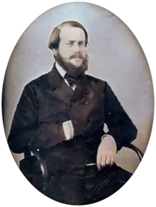 Photographie d'un jeune homme barbu assis dans un fauteuil avec sa main droite rentrée dans l'avant de sa veste à double boutonnage