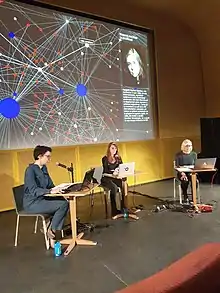 Projection d'une galaxie de liens en arrière plan, 3 femmes réparties sur une scène lisant du texte à tours de rôle sur leur ordinateur portable, dans un micro