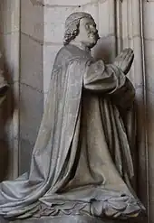 Personnage habillé d'un manteau et agenouillé, les mains jointes, tourné vers la droite.