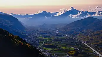 Paysage montagneux d'une large vallée vu de haut durant un coucher de soleil. On y voit plusieurs villages, des champs et un fleuve. Au loin, les montagnes sont dans les nuages.