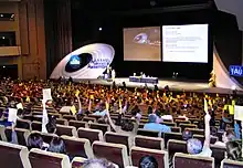 Photographie d'un amphithéâtre où de nombreuses personnes votent en levant la main avec un carton coloré.