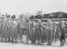 Photo en noir et blanc figurant des hommes portant sur leurs épaules un tronc passé au travers d'une monnaie de pierre