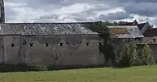 Photographies en couleurs d'un bâtiment oblong couvert d'une toiture en ardoise et percé de petites baies carrées, une étendue herbeuse le longeant au premier plan.