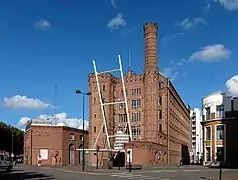 Photographie en couleur représentant un bâtiment industriel en brique rouge de cinq étages vu en enfilade, flanqué d'une tour crénelée à gauche et d'une tour plus étroite prolongée en cheminée, elle aussi crénelée, à droite